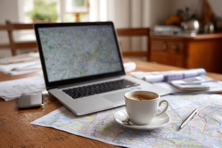 A cozy desk setup features a laptop displaying maps alongside a cup of coffee and printed maps creating an inviting atmosphere for work or planning.の素材