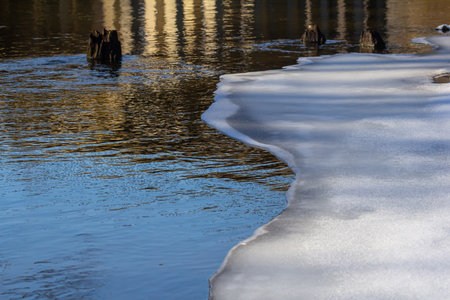 A peaceful riverside showcases melting ice along the bank reflecting sunlight while the calm water...