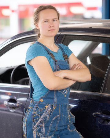 young female mechanic blue overalls stands confidently with her arms crossed next to a car in a workshop showing her skills in auto repair.の素材