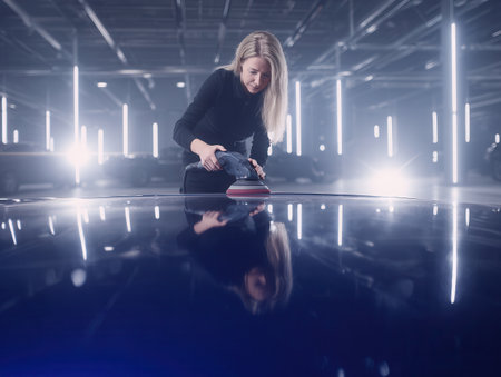 female mechanic is meticulously polishing the surface sleek blue sports car in a spacious garage with bright lights showing her craftsmanship during the evening.の素材
