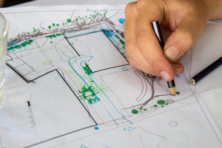 A hand holds a pencil sketching details on a landscape design showing garden elements like paths...