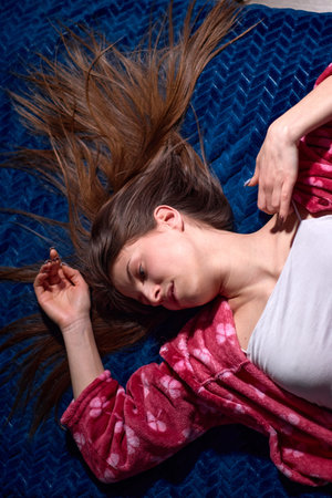 Overhead shot of a young woman with long brown hair lying on a dark blue textured blanket in soft morning light, wearing a white top and pink floral robe.の写真素材
