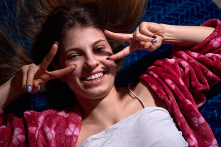A smiling teenage girl wearing a pink bathrobe and a white t-shirt is lying on a blue blanket and...