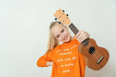 Teenage, blond girl in blue T-shirt is holding light brown ukulele instrument.