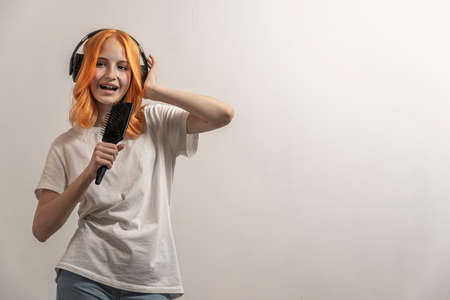 portrait of a teenage girl with red hair and in a white T-shirt singing in a hairbrush on a light...
