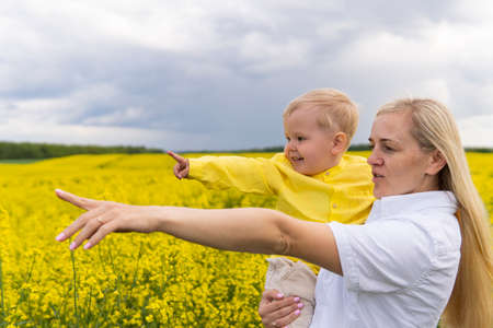 mom and son with blond hair on the field of yellow flowers point into the distance.の写真素材