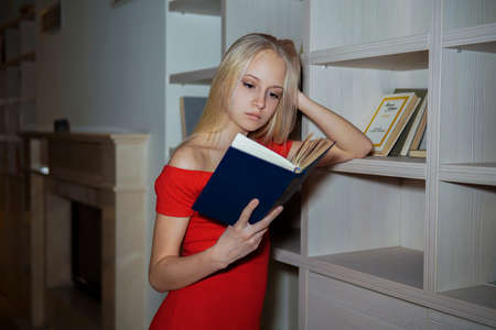 little girl teenager blonde in a red dress reading books on the background of bookshelvesの写真素材