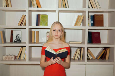 little girl teenager blonde in a red dress reading books on the background of bookshelvesの写真素材