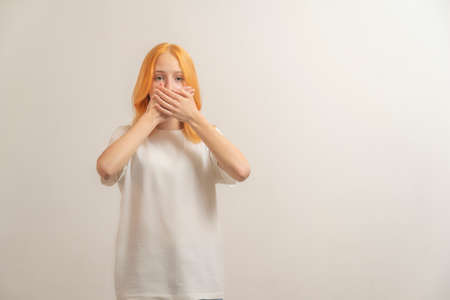 teen girl with red hair in a white t-shirt on a light background cover her mouth with her hands.の写真素材