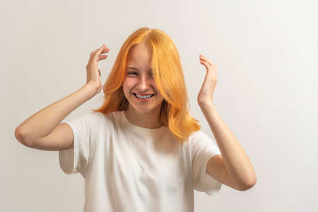teen girl with red hair in a white t-shirt on a light background laugh and straighten hair.の写真素材