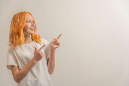 teen girl with red hair in a white t-shirt on a light background laugh and point fingers to the side.の写真素材
