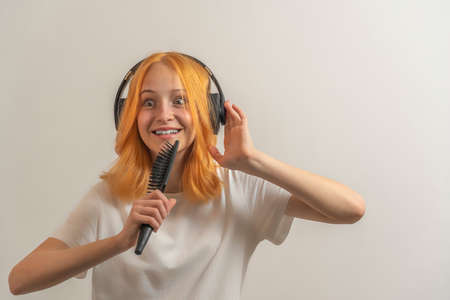 teen girl with red hair in a white t-shirt on a light background listening to music with headphones and singing.の写真素材
