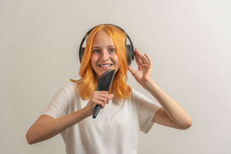 teen girl with red hair in a white t-shirt on a light background listening to music with headphones...