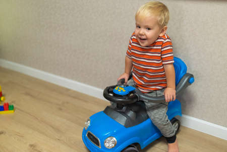 little blond boy sitting in a toy car at home.