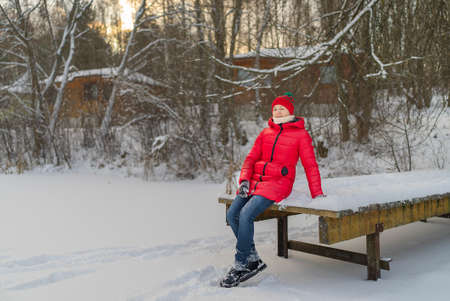 a blonde woman in a red jacket sit on a wooden pier on a snow-covered lake winter coldの写真素材
