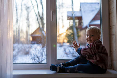 little boy stand near the window and look at the snowy street in winter, when it is very coldの写真素材