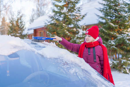 young woman in a red hat cleaning a snow-covered car on the background of a winter road and the setting sunの写真素材