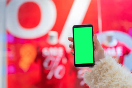 woman's hand holds a phone with a green screen to replace the background, in the mall with the background of a shop window with discounts during a sale.の写真素材