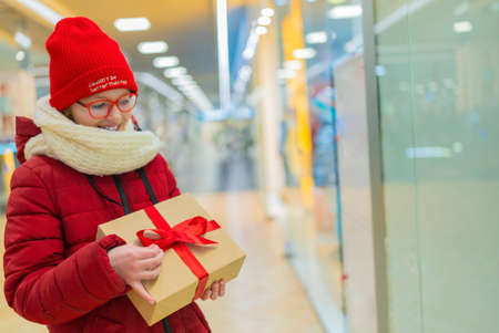 teenage girl in a white jacket in a mall, holding a box with a gift on the background of a shop window with discounts during a sale.の写真素材