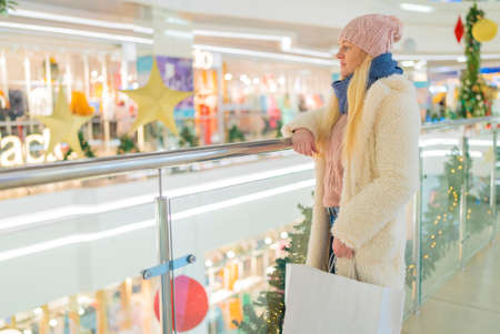 a teenage girl in a white jacket in a shopping center, speaks on the phone against the background of a shop window with discounts during a sale.の写真素材