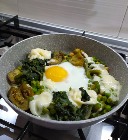 Fried eggs with vegetables in a frying pan on a gas stove, home cooking. Scrambled eggs with spinach, eggplant, peas, beans, close-up.の写真素材
