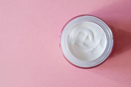An open glass jar with white cream for the face or body on a pink background. Skincare concept....