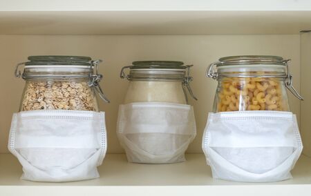 Glass jars with stocks of products in masks stand at a distance from each other on a shelf in a cupboard. Social distancing, wearing a mask, COVID-19 concept. Horizontal orientation, selective focusの写真素材