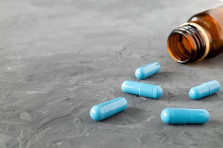 The blue-colored medical capsules woke up from an overturned glass bottle that lays on a gray...