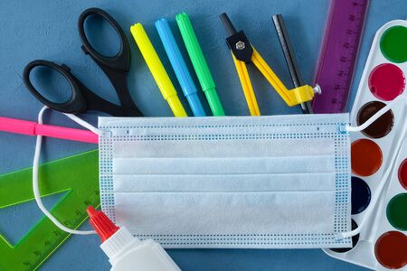 School supplies and medical mask on a blue background. Back to school, the pandemic coronavirus concept. Horizontal orientation. Selective focus. View from above.の写真素材
