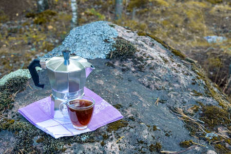 A geyser coffee maker and a small glass cup of coffee on a linen napkin stand on a large stone in...