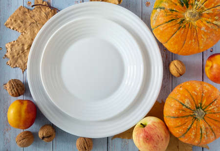 White empty porcelain plates of pumpkin, walnuts, apple and nectarines with autumn leaves on a blue background. Horizontal orientation, selective focus, top view. Mocup.の写真素材