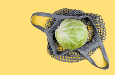 A head of cabbage lies in an ecological string bag on a yellow background. Eco friendly shopping, environmental protection concept. Colors of the Year 2021. Illuminating yellow and ultimate gray.の写真素材