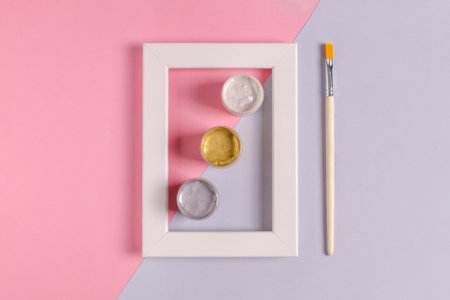 Mock up of a white blank frame with cans of three colors of acrylic paint and a brush for decoration and painting. Pink and lilac background, minimalism. Selective focus.の写真素材