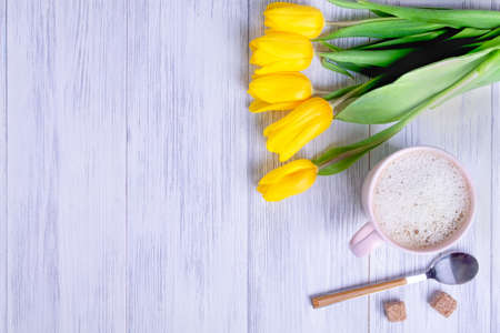 Top view of a composition of a bouquet of yellow tulips, a pink cup with cappuccino with a spoon and sugar on a light wood background. Selective focus. Copy space for your text.の写真素材