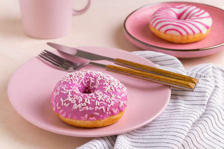 Two sugar-coated donuts lie on a pink ceramic plates. Serving with cutlery, mug and linen napkin. Selective focus.の写真素材