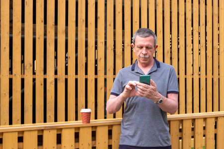 An adult pensioner in a gray T-shirt stands against the background of a wooden wall and touches the smartphone screen. Selective focus.の写真素材