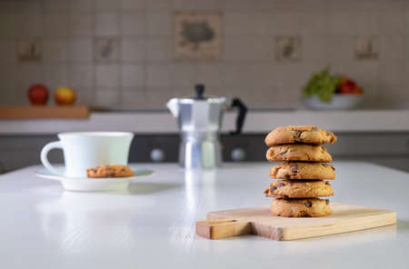 Stack of oatmeal cookies with chunks of chocolate lies on table on small serving board. Morning coffee is served in cozy kitchen. Focus on foreground. Blurred focus and backdrop.の写真素材