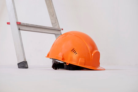 Bright orange construction helmet is lying next to stepladder against plaster wall. Renovation...