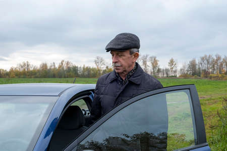 Adult retired man gets into car against backdrop of rural landscape in autumn. Country life....