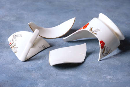 Close-up of fragments of white fragile porcelain cup on blue surface with stucco texture. Selective...
