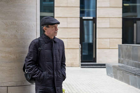 Adult Caucasian male stands near modern building with columns. Pensioner in seasonal clothes while walking around the city. Selective focus.の写真素材
