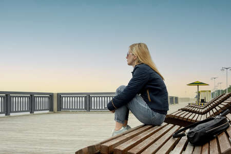 A young lonely blonde woman on an empty city embankment rests sitting on a sun lounger on a spring or autumn evening and looks into the distance. The girl misses the warmth and summer.の写真素材