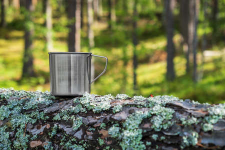 Metal tourist mug with hot drink stands on tree trunk in the forest. Tourism, outdoor activities. Selective focus.の写真素材