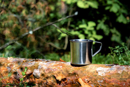 Metal tourist mug with hot drink stands on tree trunk in the forest. Tourism, outdoor activities. Selective focus.の写真素材