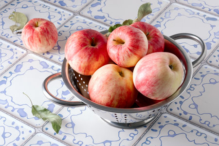 Ripe red apples lie in a metal colander on a table with a blue and white tile surface. Selective focus.の写真素材