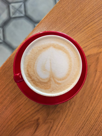 Cappuccino cup featuring heart-shaped latte art in a vibrant red cup. The coffee sits on a wooden table, highlighting the intricate design created in the creamy foam.の写真素材
