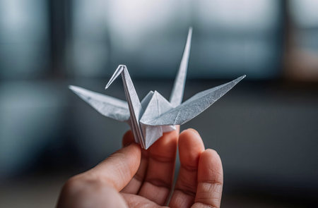 Hand holds a delicate origami crane against a blurred background, showcasing art and craftsmanship. Cultural significance of origami as a symbol of peace and creativity.の素材