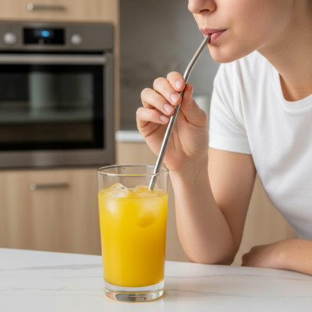 Woman savoring a refreshing glass of orange juice through a metal straw in a modern kitchen. AI generated.の素材