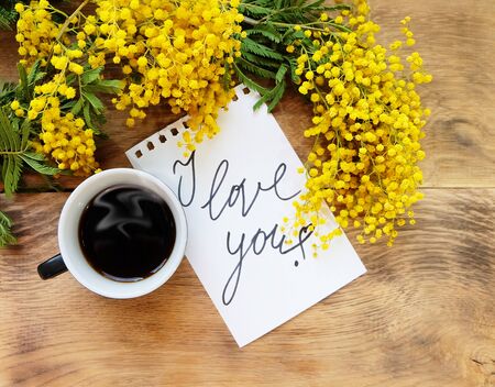 A cup of black coffee, a mimosa and a sheet of notebook with the inscription "I love you!" Spring still life with mimosa on a wooden surface.の写真素材