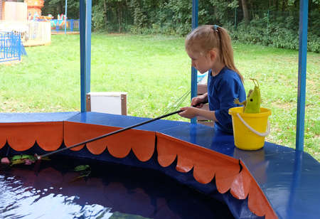 A little girl with blond long hair tied up in a ponytail is catching fish at a children's "fishing" attraction.の写真素材